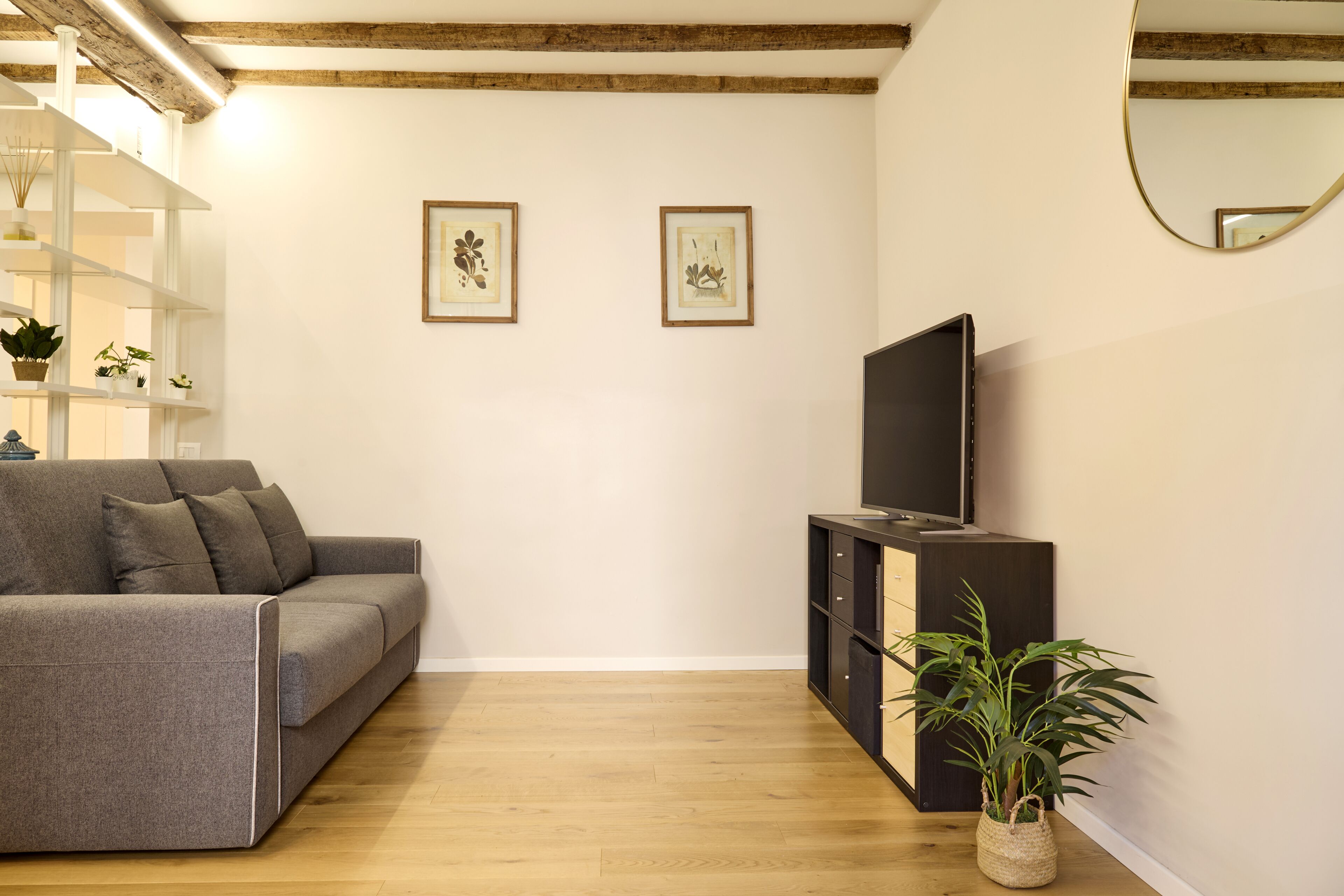 Una sala de estar con un sofá gris, un televisor sobre un mueble negro, suelo de madera y dos cuadros en la pared.