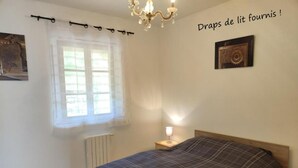 Ein Schlafzimmer mit einem Bett, einem Fenster mit durchsichtigen Vorhängen, einem Heizkörper, einer Nachttischlampe und einer Deckenleuchte.