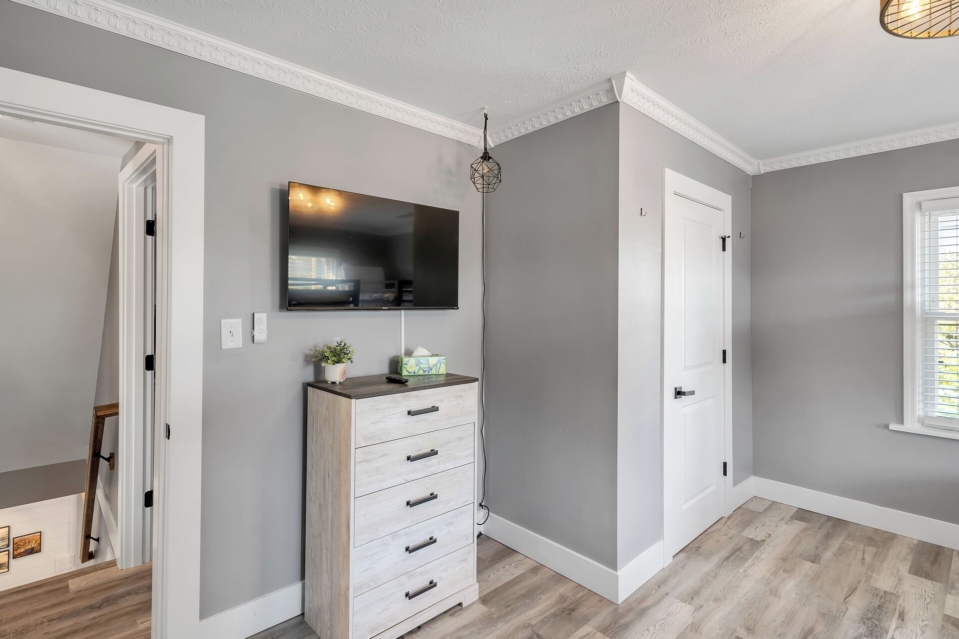 Un couloir avec un plancher en bois, un mur blanc, un téléviseur fixé au mur, une porte blanche et une fenêtre avec des stores.