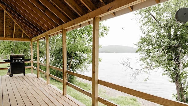 A covered wooden deck with a grill and a view of a lake. - Portage Lakeside Cabins (Portage Lake)