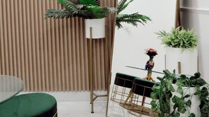 Una habitación moderna con un espejo grande, un taburete acolchado verde, una mesa redonda y varias plantas en macetas.