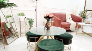Una mesa redonda de cristal con taburetes tapizados en verde, un sofá rosa y un jarrón con flores.