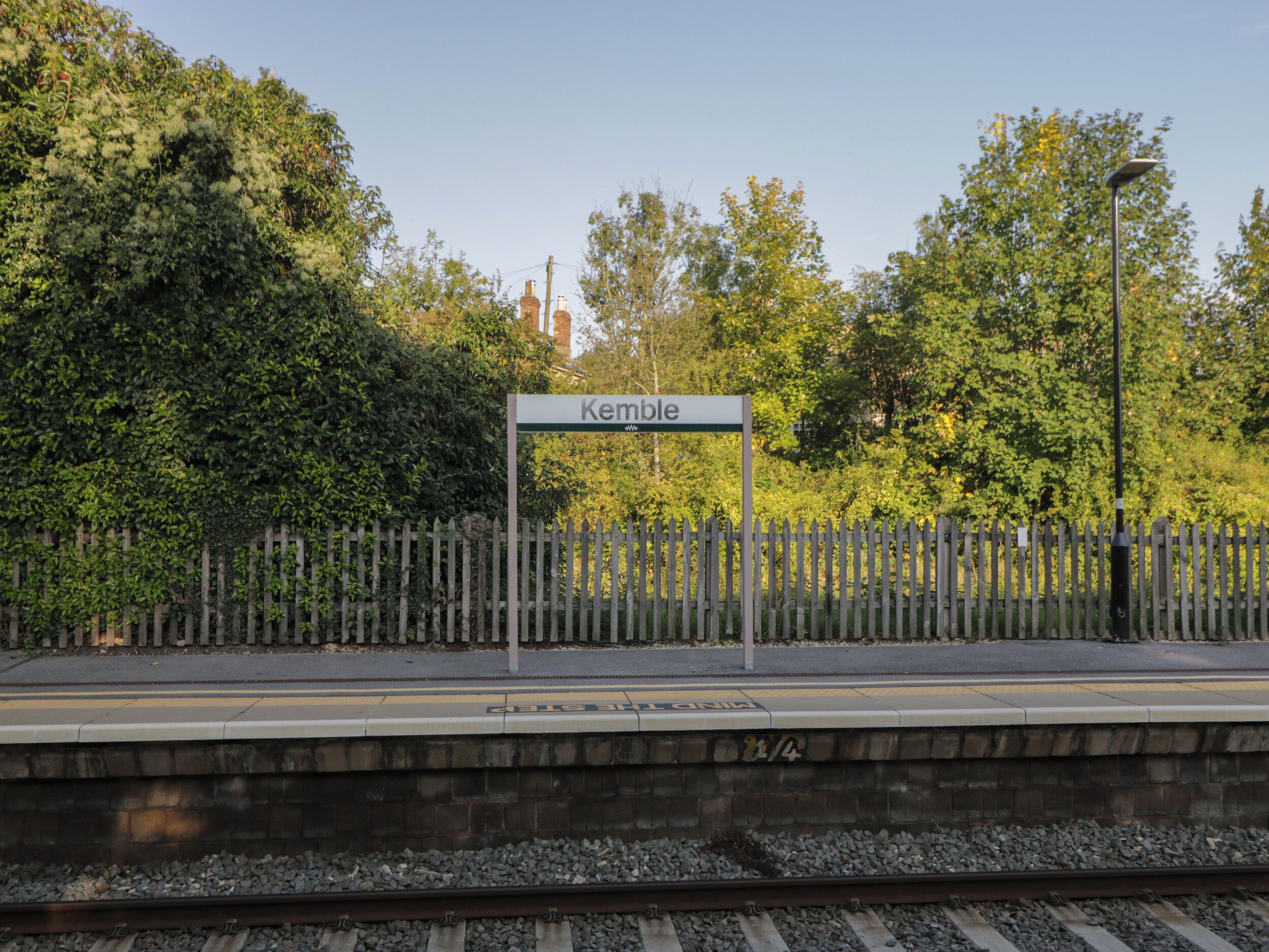 A train station platform with a sign reading "Kemble" and a wooden fence in the background.