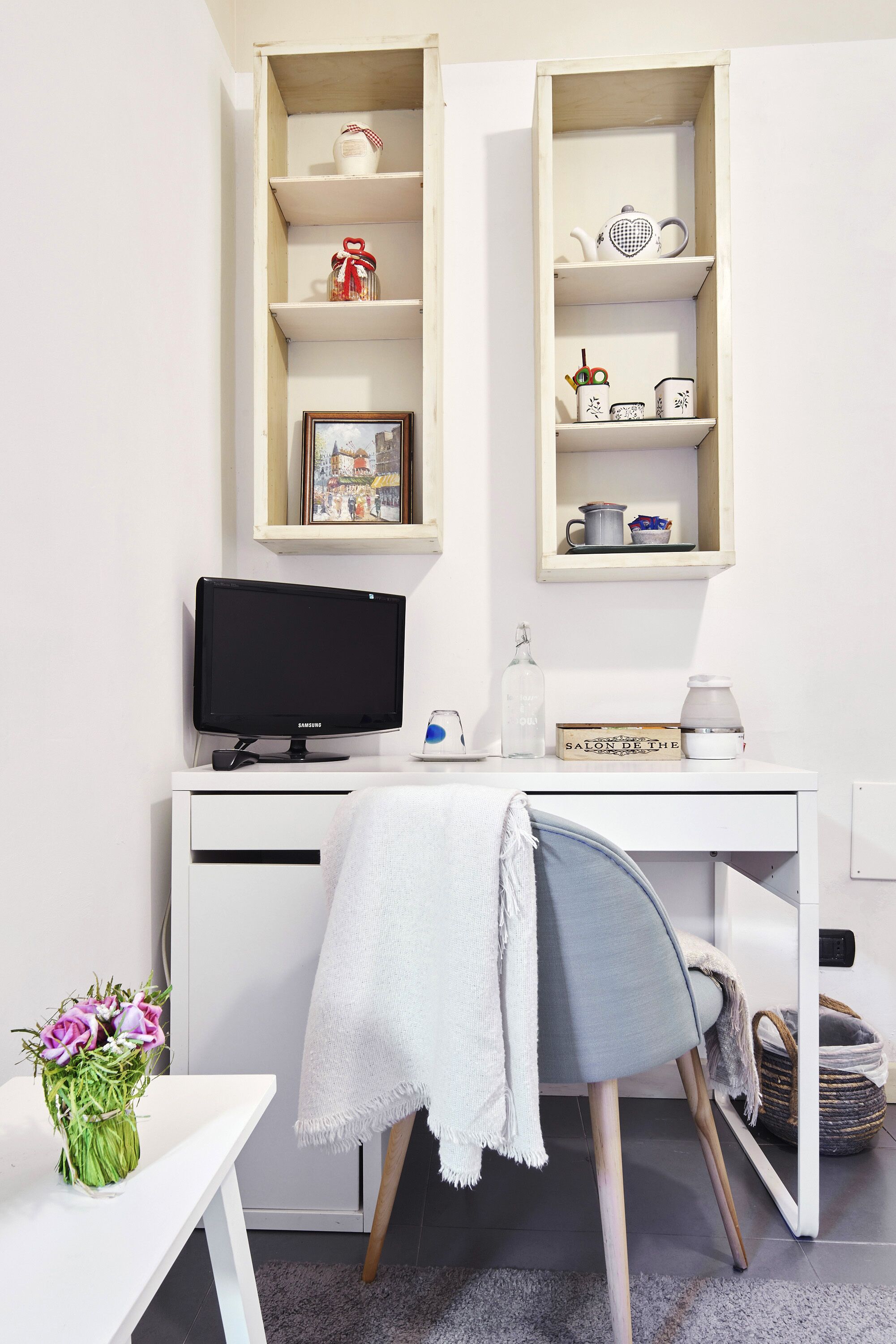 Una habitación moderna y minimalista con un escritorio blanco, un monitor de computadora, una estantería con objetos decorativos y una pequeña mesa con un florero.