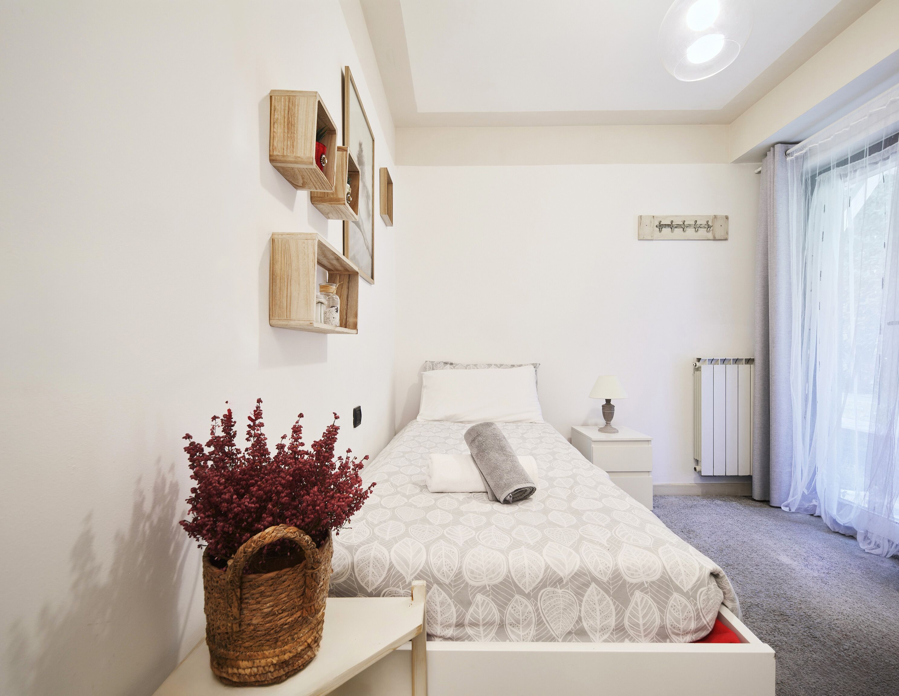 Un dormitorio con cama, mesita de noche, estantes en la pared, una ventana con cortinas y una planta en maceta.