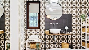 Ein Badezimmer mit Wand in geometrischem Muster, einem runden Spiegel, einem weißen Waschbecken und einem kleinen Fenster.