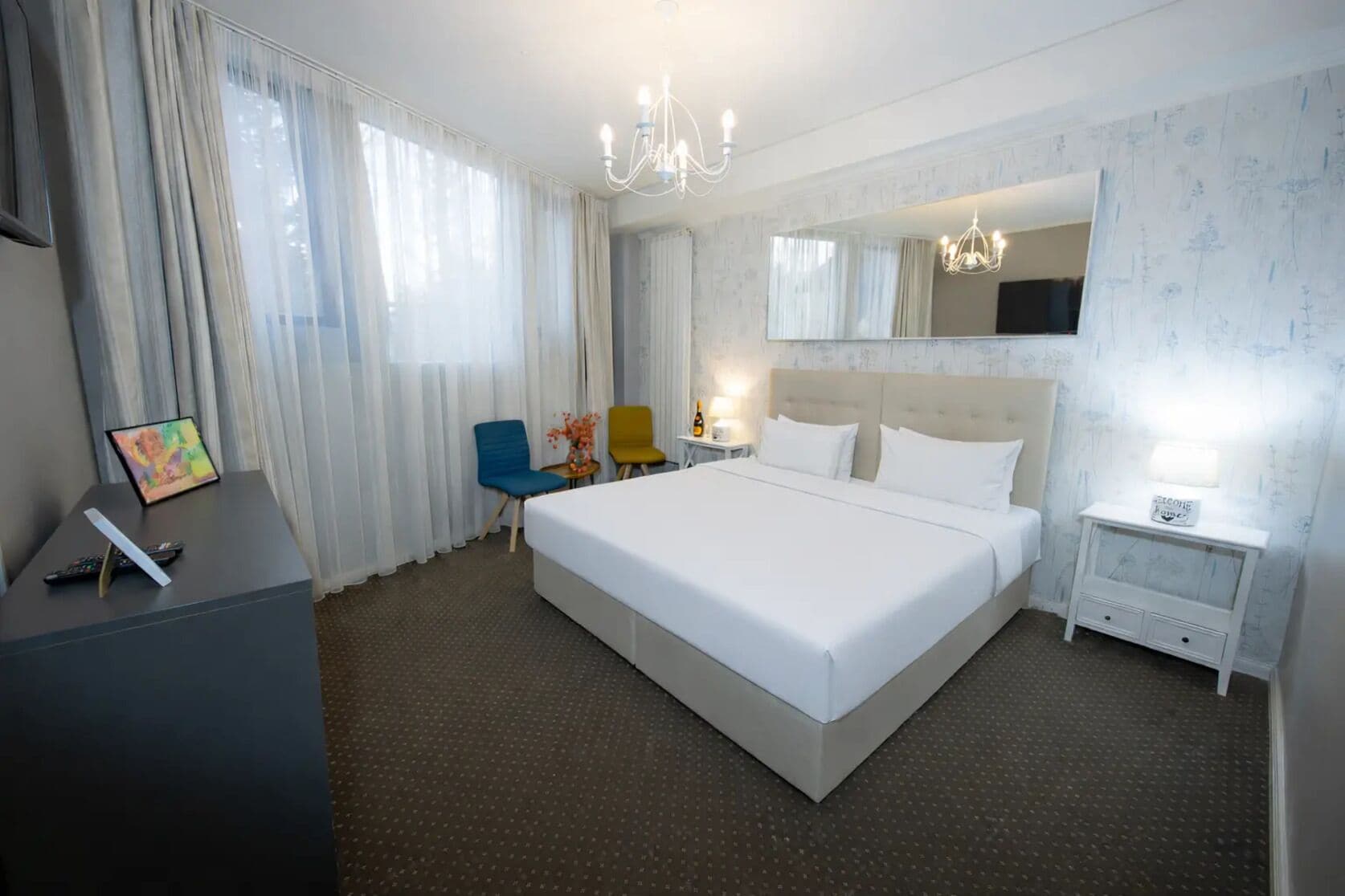 Een hotelkamer met een groot bed, een bureau met stoel, een kleine tafel met lamp, en een spiegel aan de muur.