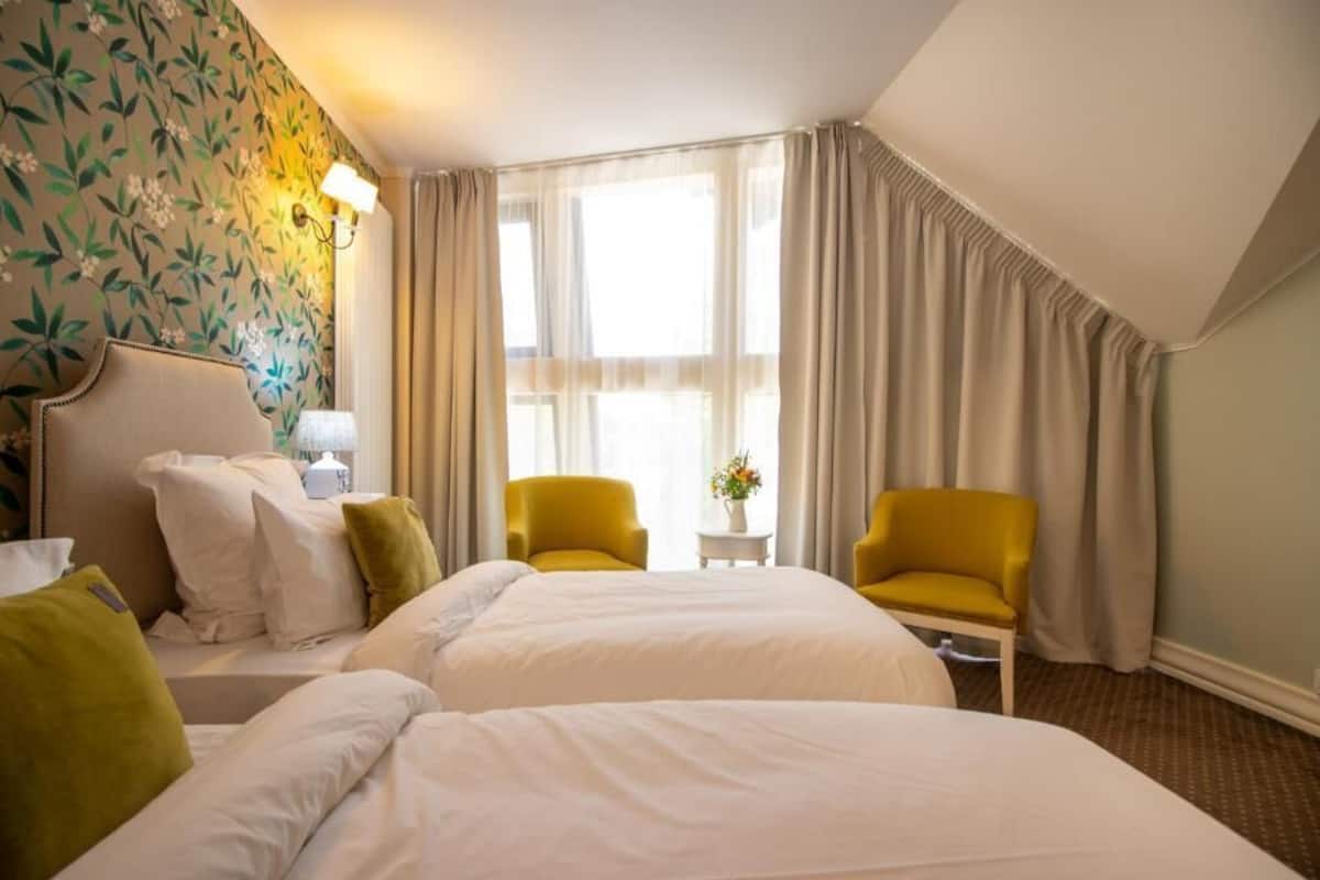 Een hotelkamer met twee bedden, een nachtkastje, een lamp, een kleine tafel met een vaas, twee stoelen en een raam met gordijnen.