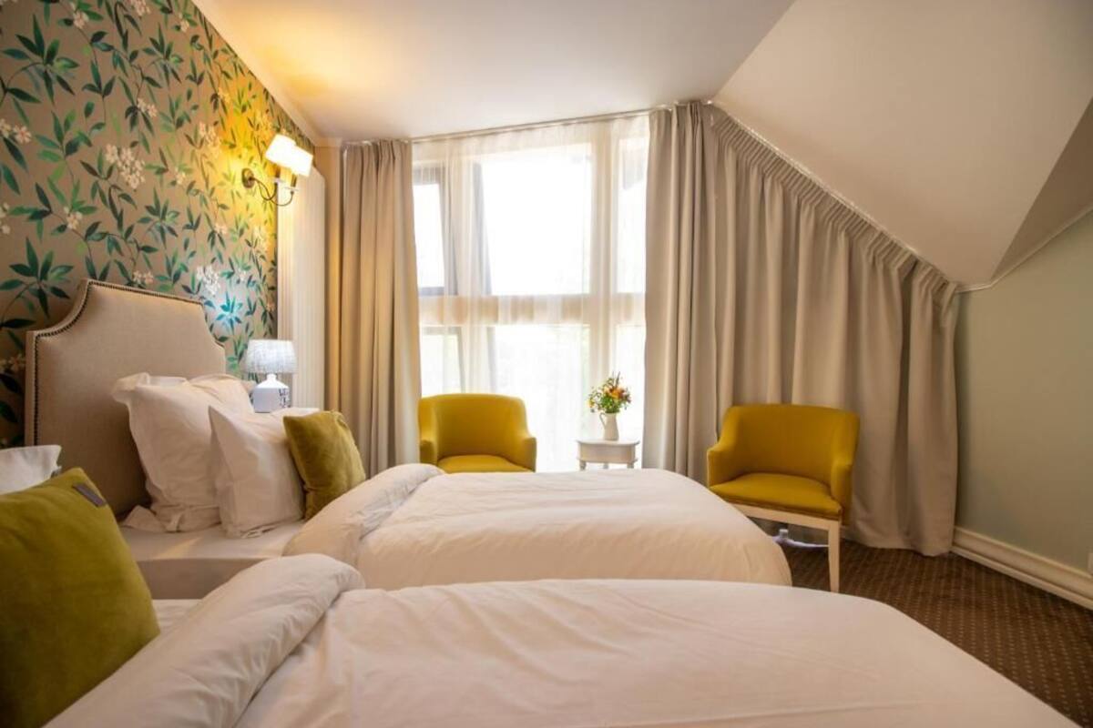 Ein Hotelzimmer mit zwei Betten, einem Nachttisch, einer Lampe, einem kleinen Tisch mit einer Vase, zwei Stühlen und einem Fenster mit Vorhängen.