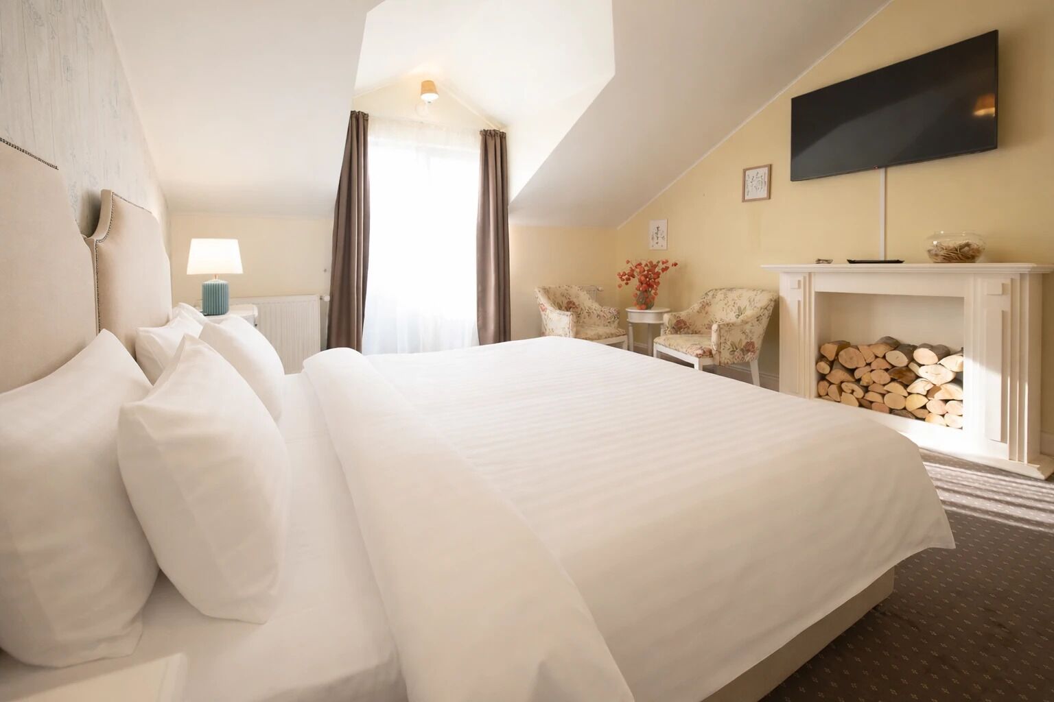 A hotel room with a large bed, a flat-screen TV mounted on the wall, a fireplace with logs, and two chairs.