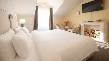 A hotel room with a large bed, a flat-screen TV mounted on the wall, a fireplace with logs, and two chairs.