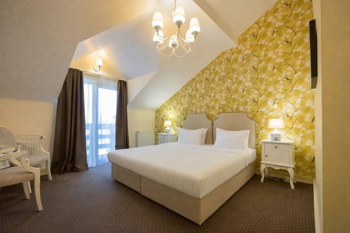 Een slaapkamer met een groot bed, een stoel, een nachtkastje, een kroonluchter en een raam met gordijnen.