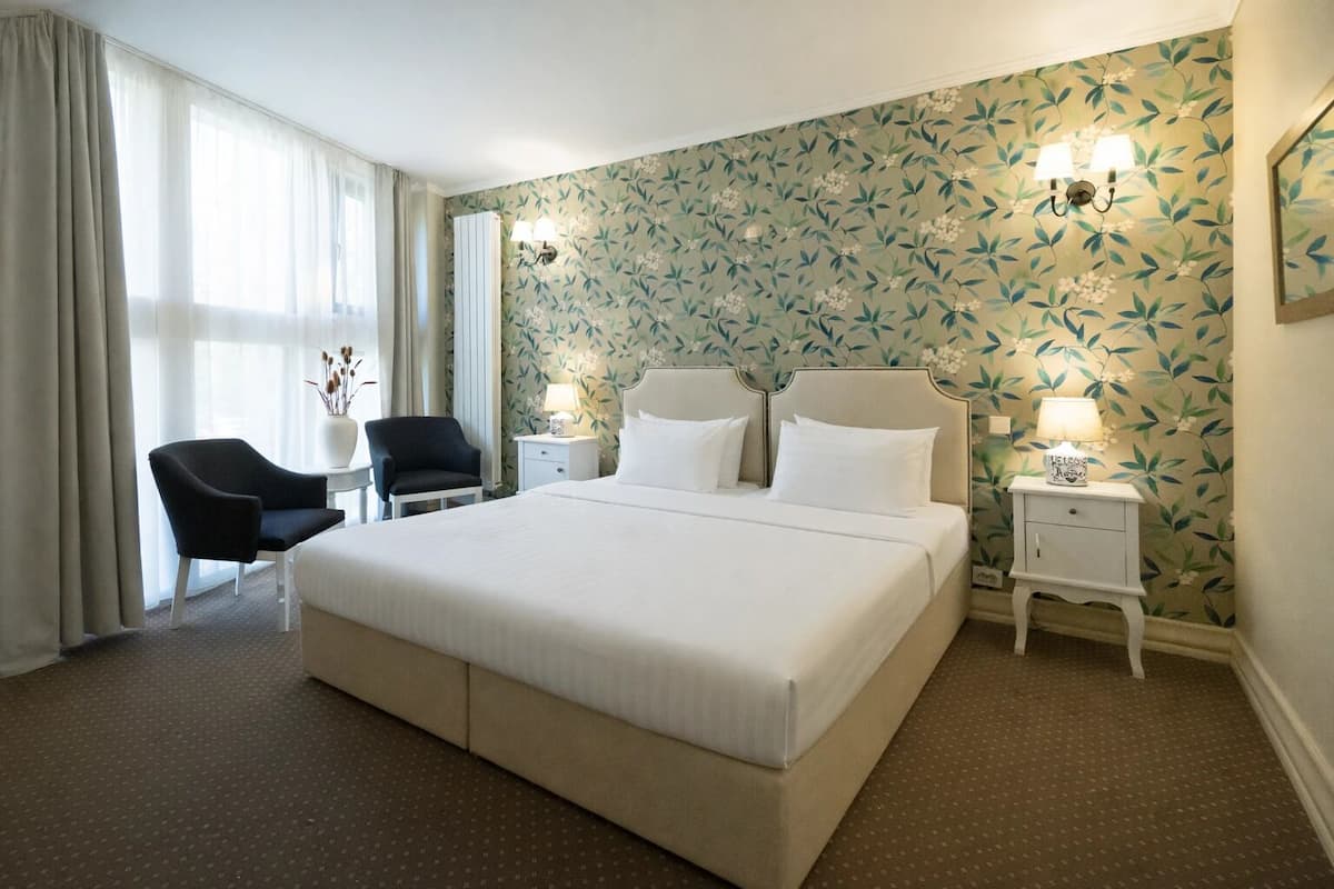 Een hotelkamer met een groot bed, twee nachtkastjes, een stoel, een klein tafeltje en een raam met gordijnen.