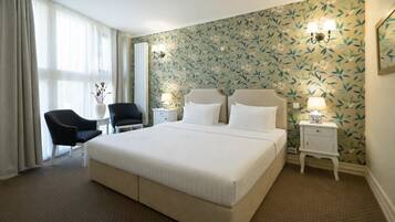 A hotel room with a large bed, two bedside tables, a chair, a small table, and a window with curtains.