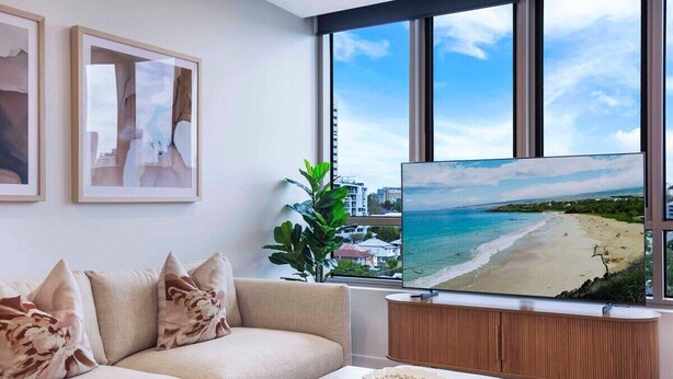 Un salon moderne doté d’un grand téléviseur diffusant une vue sur une plage, d’un canapé beige et d’une console en bois.