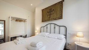 A neatly made bed with white linens and towels, a white wardrobe, a bedside table with a lamp, and a tapestry on the wall. - Poliziano Apartment by Mmega (Florence)