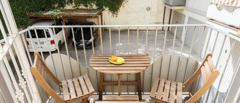 A balcony with a wooden table and two chairs, a white car parked in the background, and a green wall with climbing plants.