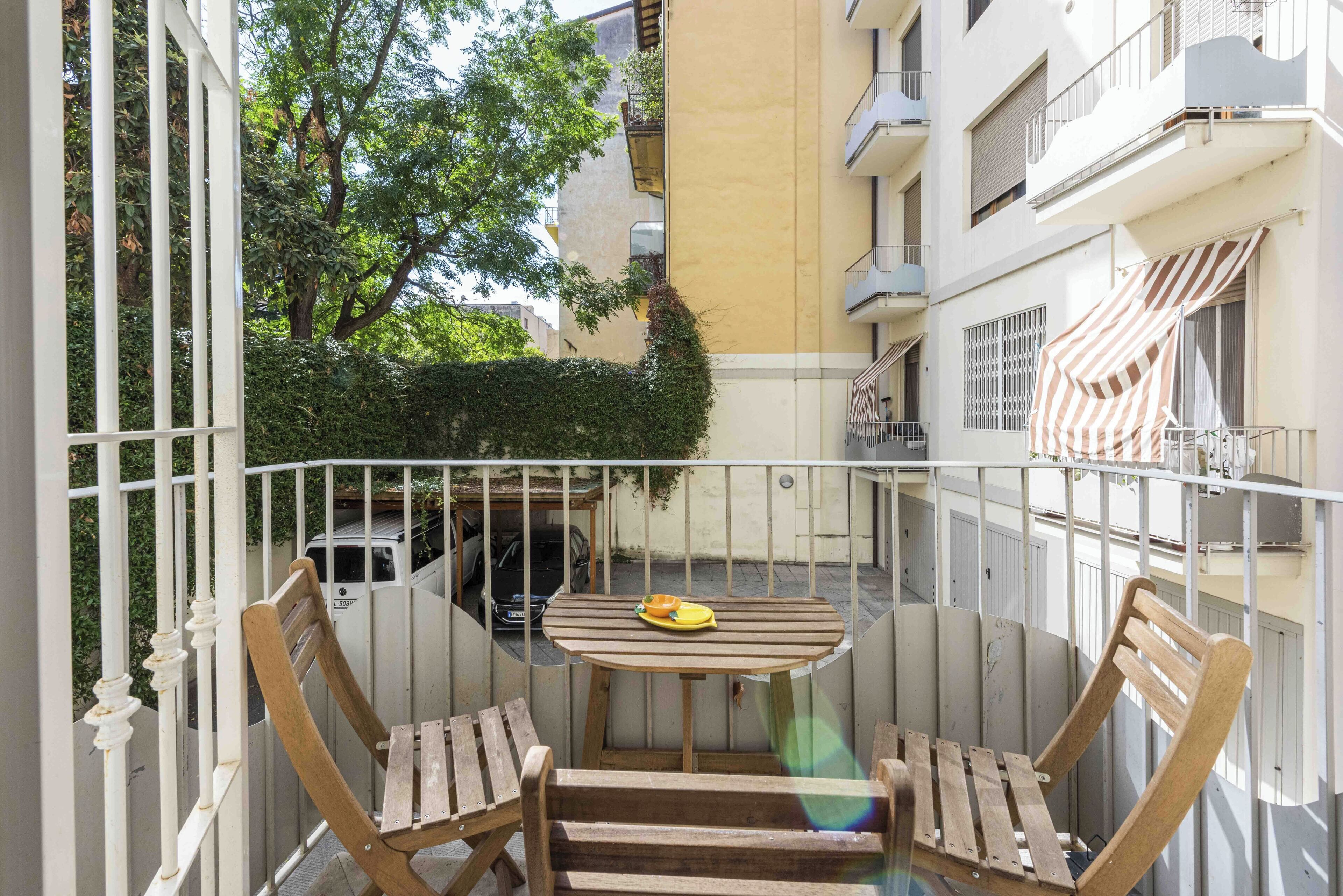 A balcony with a wooden table and chairs, a yellow bowl, and a white car parked in the background.