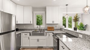 A modern kitchen with white cabinets, granite countertops, stainless steel appliances, and a large window with a view of greenery. - Cape Fear Treehouse Boat Ramp, Dock, Kayaks, Firepit, Games (Pender County)