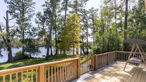 A wooden deck with a swing chair and a table, overlooking a lake surrounded by trees. - Cape Fear Treehouse Boat Ramp, Dock, Kayaks, Firepit, Games (Pender County)