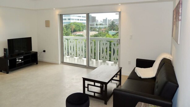 A modern living room with a flat-screen TV, a sofa, a small table, and a balcony with a view of buildings and greenery.