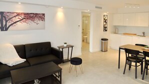 A modern living room with a black sofa, a small round table, and a painting on the wall. There is a kitchen area with white cabinets and a dining table with chairs.