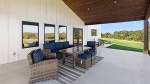 Un patio exterior con mobiliario de mimbre, una mesa de vidrio y grandes ventanales con vistas al paisaje.