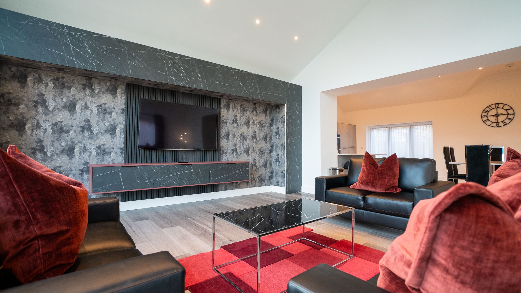A modern living room with a large flat-screen TV, dark wood flooring, and red upholstered sofas.