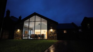 A modern house with a large glass facade, stone walls, and a visible interior with a dining area and a clock. - Northumberland Luxury Stays - The Shearling (Morpeth)