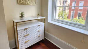 Een hoekzaak met een witte console tafel, een decoratief gouden object, een kleine plant, een kast met een herringbone-patroon, een houten vloer en uitzicht op een gebouw van rode baksteen door een raam met jaloezieën.