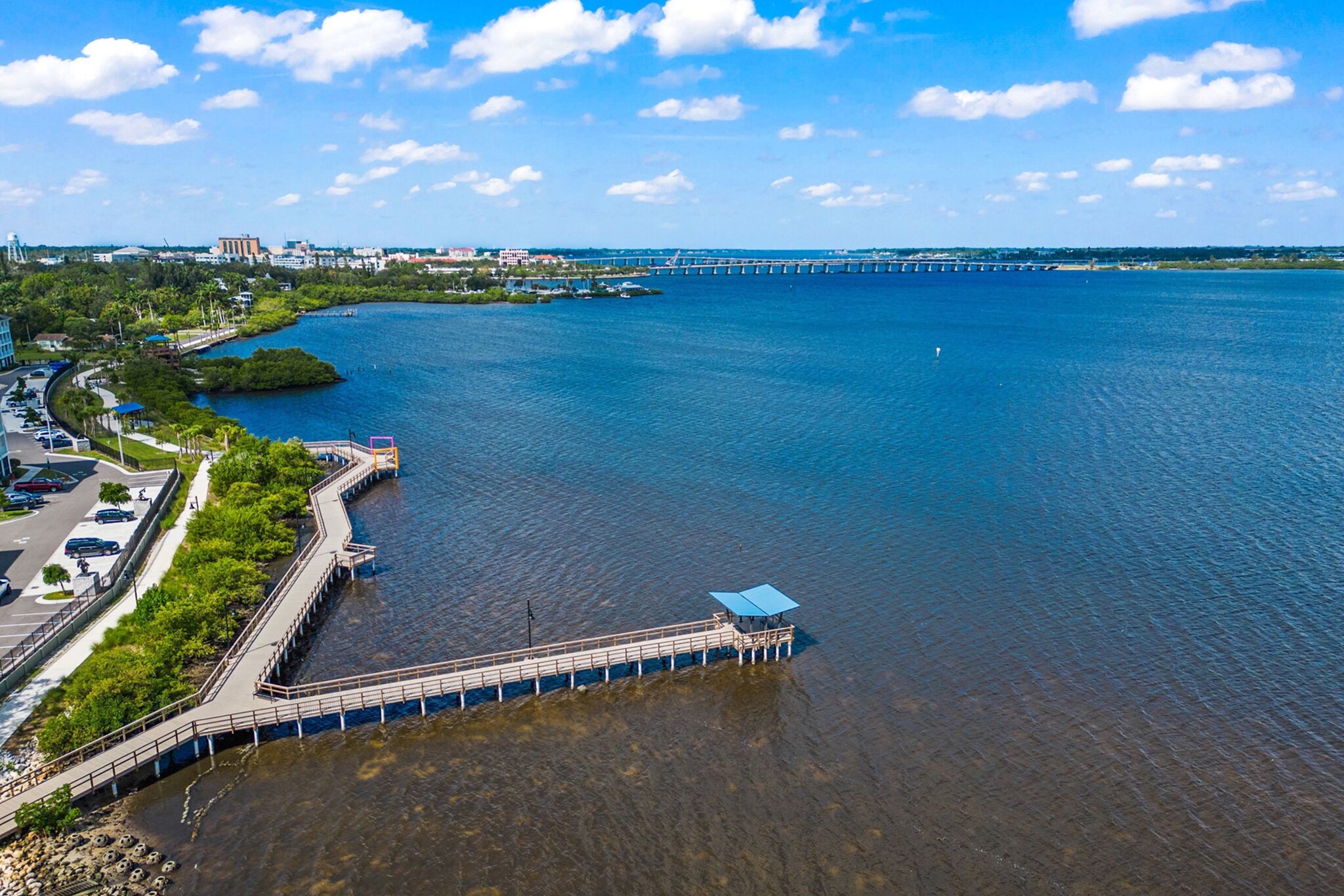 Un muelle frente al agua con una plataforma de observación, un puente al fondo y un paisaje urbano con edificios y un estacionamiento.