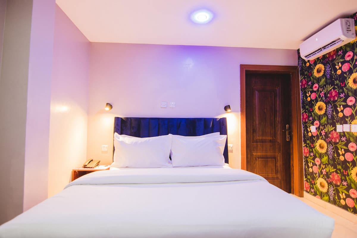 A hotel room with a bed, bedside lamps, an air conditioning unit, and a floral wallpaper.