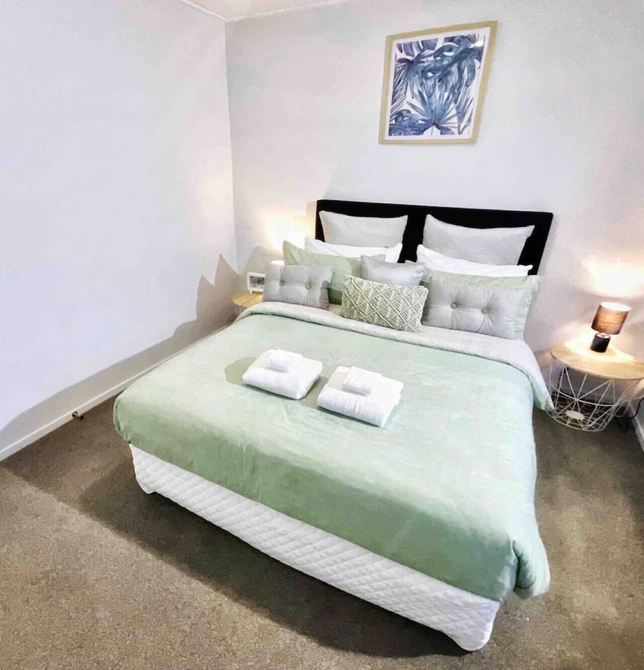 A neatly made bed with a green bedspread and white pillows, a framed artwork on the wall, and a bedside lamp.