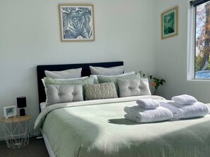 A neatly made bed with a green bedspread and white pillows, a bedside table with a lamp, and framed artwork on the wall. - Perfect On The Park! (Tauranga)