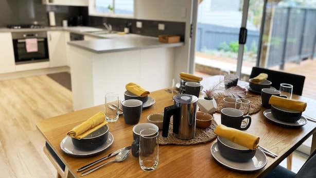 En frukostuppsättning med ett träbord, skålar, koppar och en kaffekanna i ett modernt kök med ett stort fönster.
