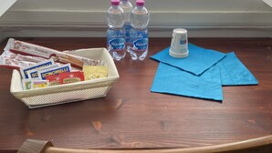 A wooden table with a basket of snacks, water bottles, and a can of soda. A window reveals a parked car and a building outside. - PROMOTION "TOURISTHOUSE MONOPOLI 10" ROOM1 TRIPLE WITH EN SUITE BATHROOM (MONOPOLI)