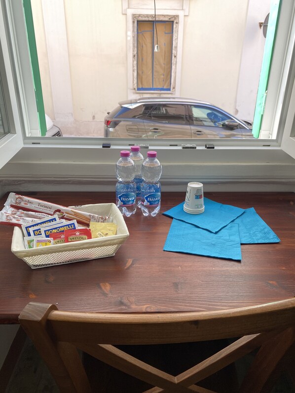 Une table en bois sur laquelle se trouve un panier de collations, des bouteilles d’eau et une canette de soda. Une fenêtre donne sur une voiture garée et un bâtiment situé à l’extérieur.