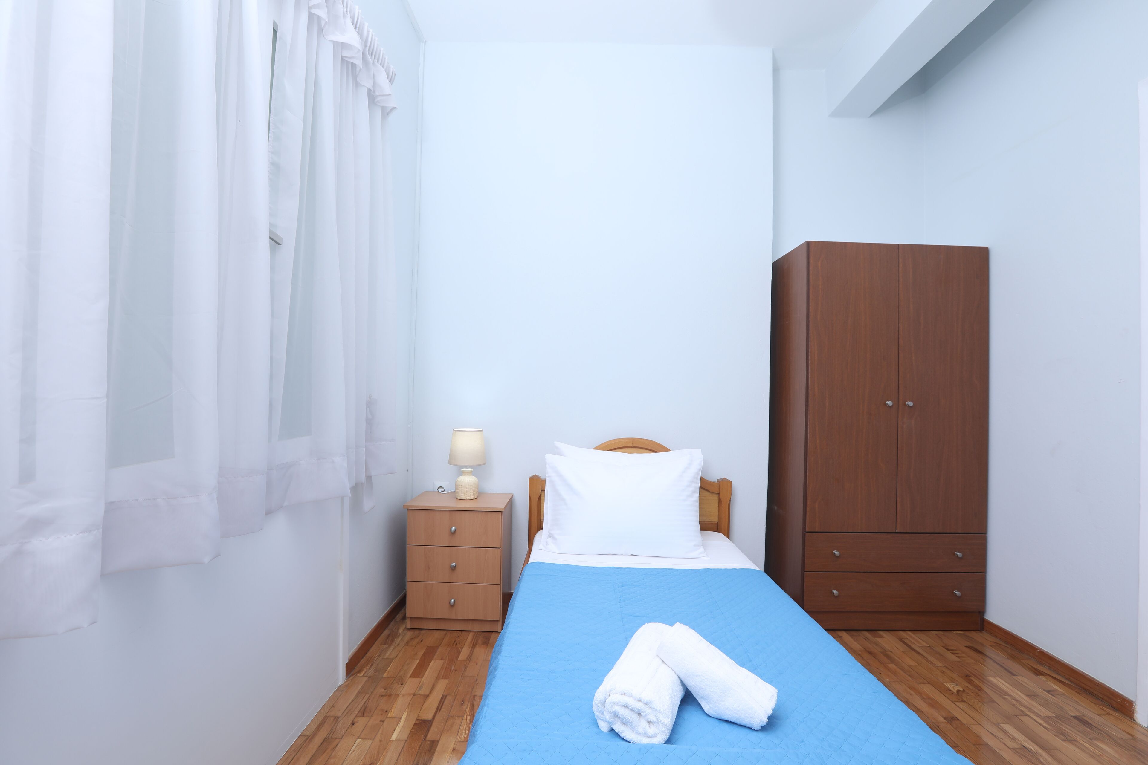 A bedroom with a bed, a nightstand, a wooden wardrobe, and a window with white curtains.