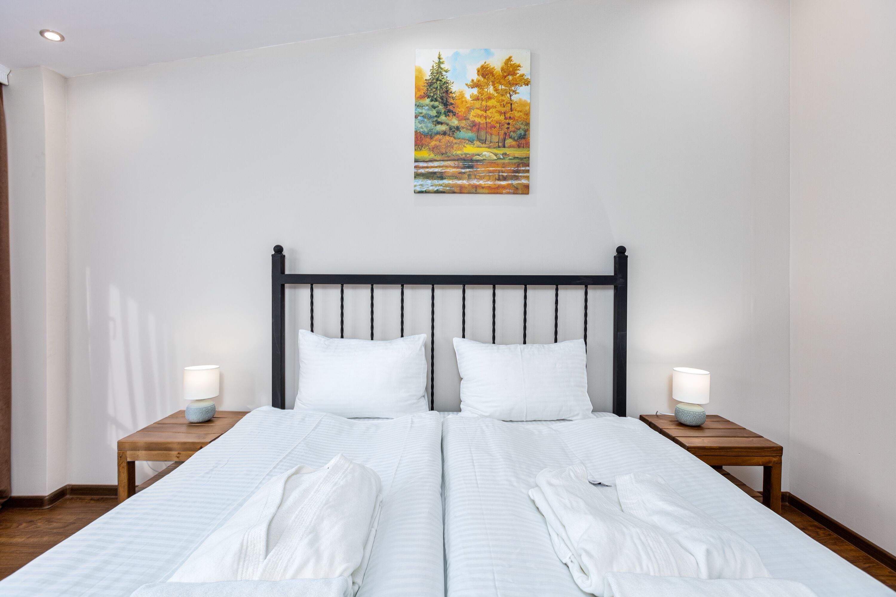 A double bed with white bedding, two bedside tables with lamps, and a painting of a landscape on the wall.