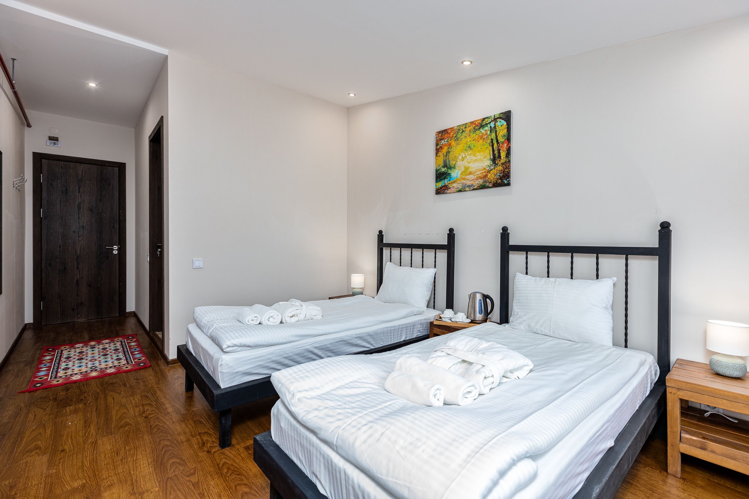 A hotel room with two beds, a wooden floor, a painting on the wall, and a small table with a lamp.