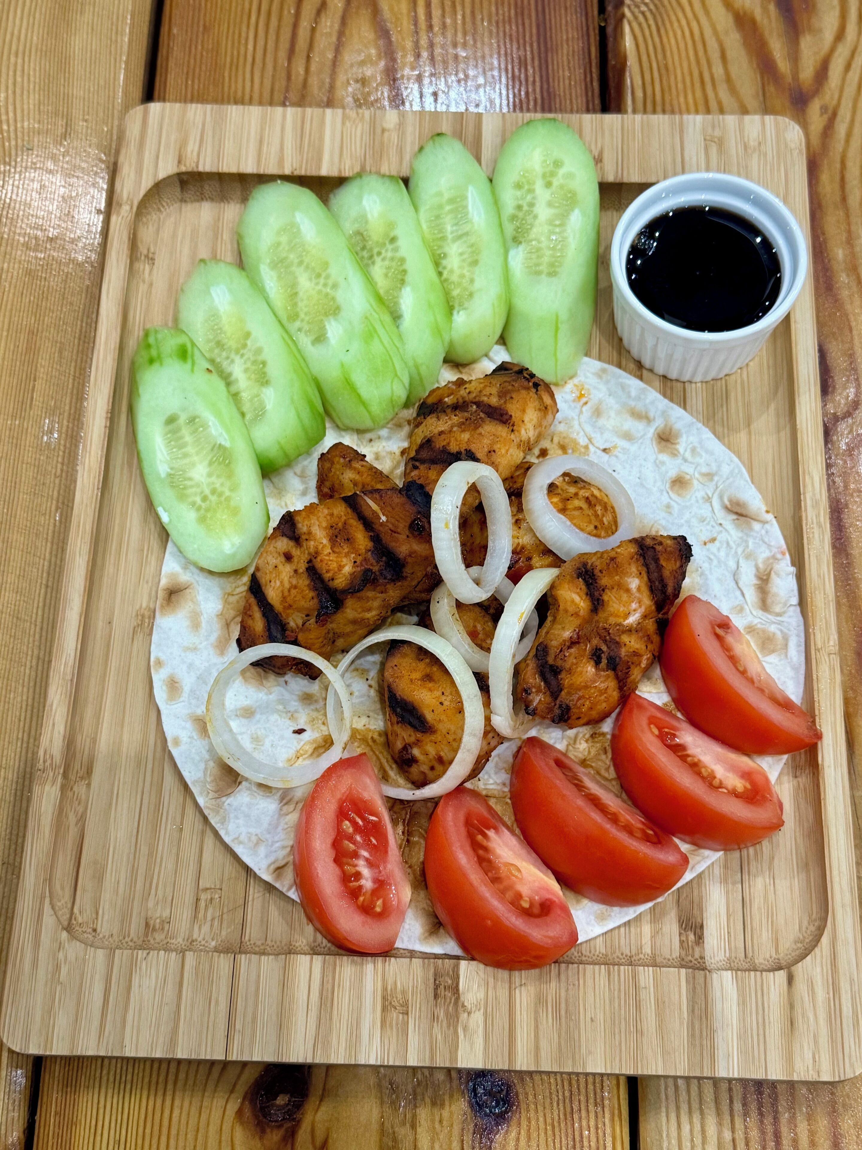A wooden cutting board with grilled chicken, sliced tomatoes, onions, and cucumbers, accompanied by a small bowl of sauce.