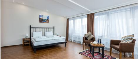 Un dormitorio moderno con cama, dos sillones, una mesita y un gran ventanal con cortinas de gasa.