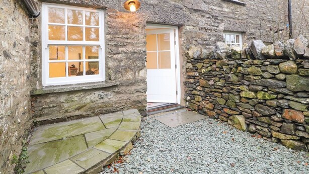 A stone building with a white door and windows, a stone wall, and a gravel path. - Hazelrigg Cottage (Ulverston)