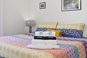 Una cama con un edredón colorido, dos almohadas con estampados florales y un juego de toallas con la marca 'MAKEUP' y 'MAKFUP' visible.