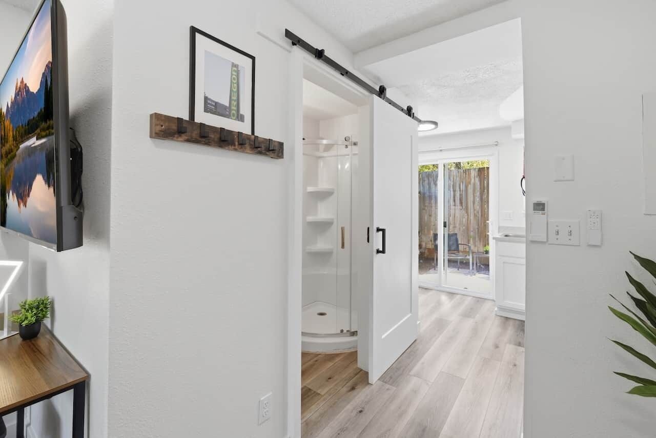 A modern interior with a mounted TV, a wooden shelf with a framed picture, a glass shower enclosure, and a view through an open door to a balcony.