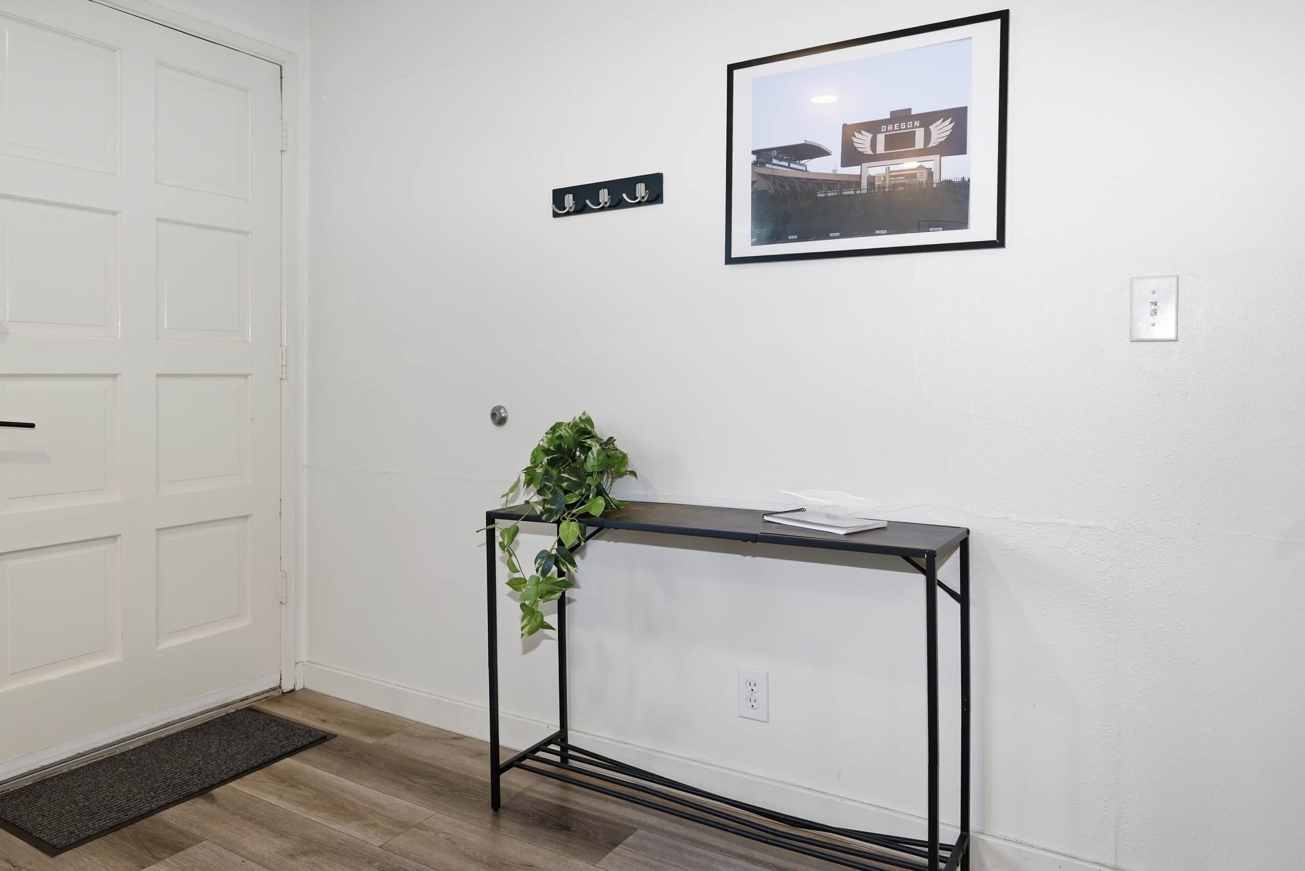 Un couloir avec une porte blanche, un tableau encadré accroché au mur, une petite table avec une plante et un paillasson.