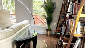 A modern living room with a white sofa, a glass coffee table, a wooden bookshelf, and a large window with a view of a red staircase.