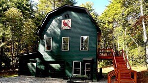 A green wooden cabin with a red staircase leading to a deck, surrounded by trees.