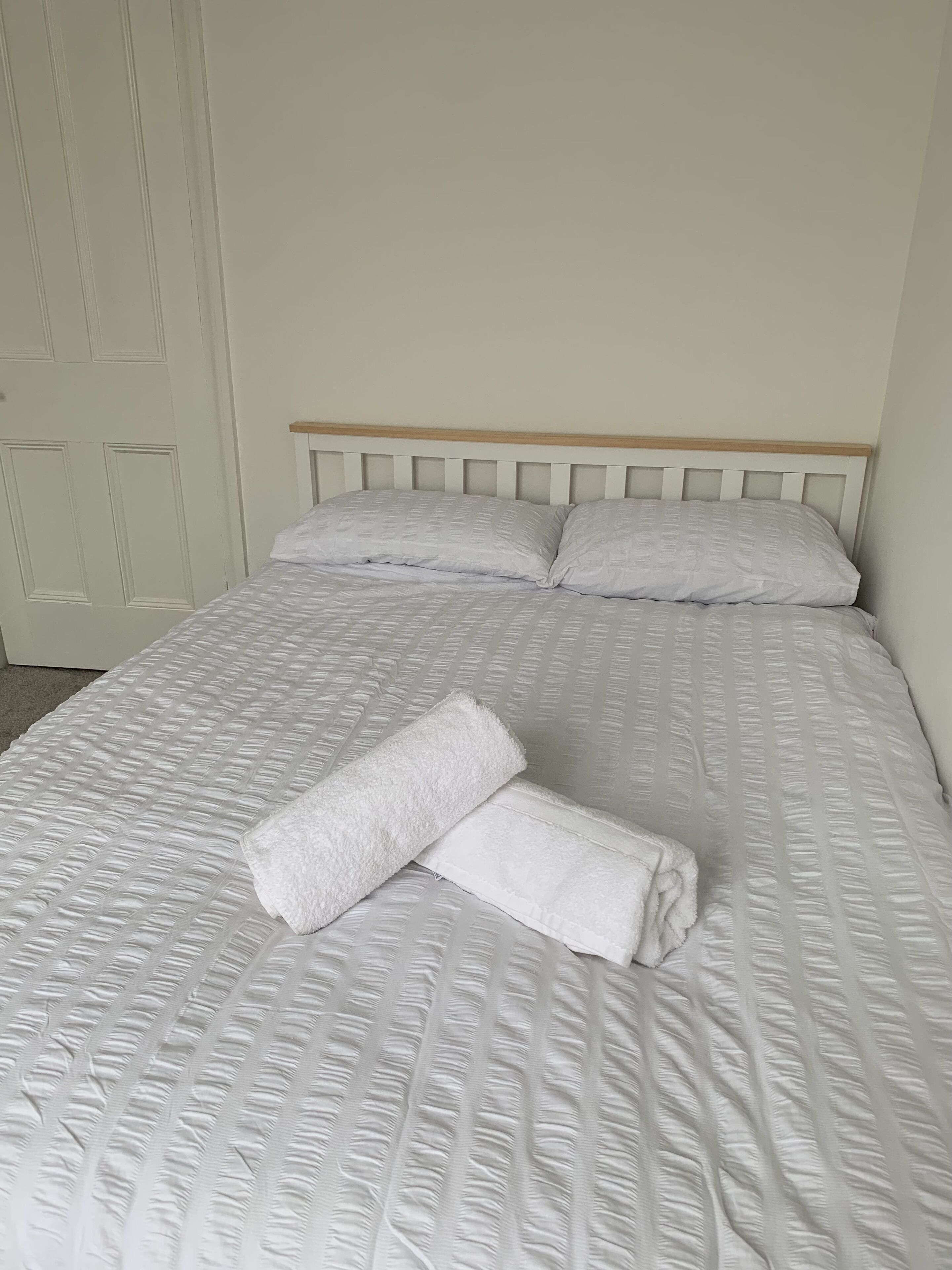A neatly made bed with a white duvet and pillows, a wooden headboard, and a white door in the background.