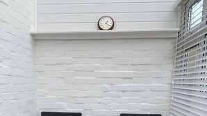 Un rincón con asientos, dos sillas de mimbre y una pequeña mesa, paredes de ladrillo blanco y un reloj en la pared.